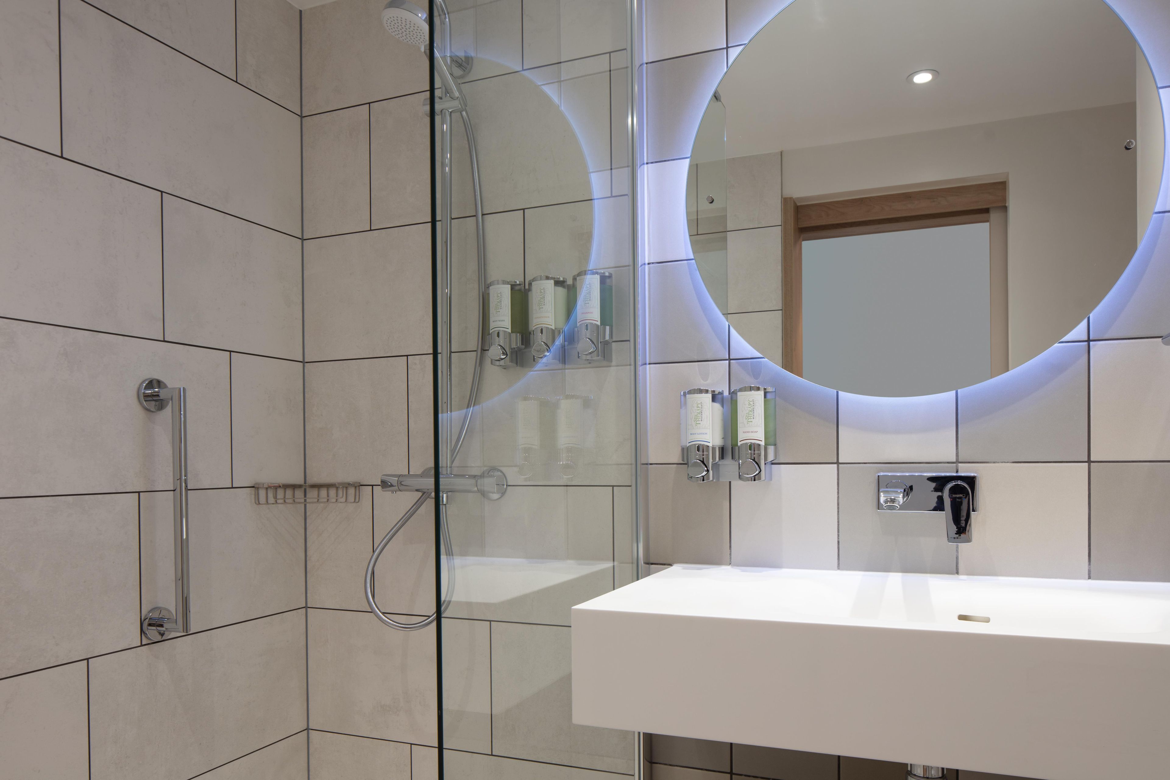 Un baño moderno con cabina de ducha de vidrio, un lavabo blanco y un espejo con luz circular.