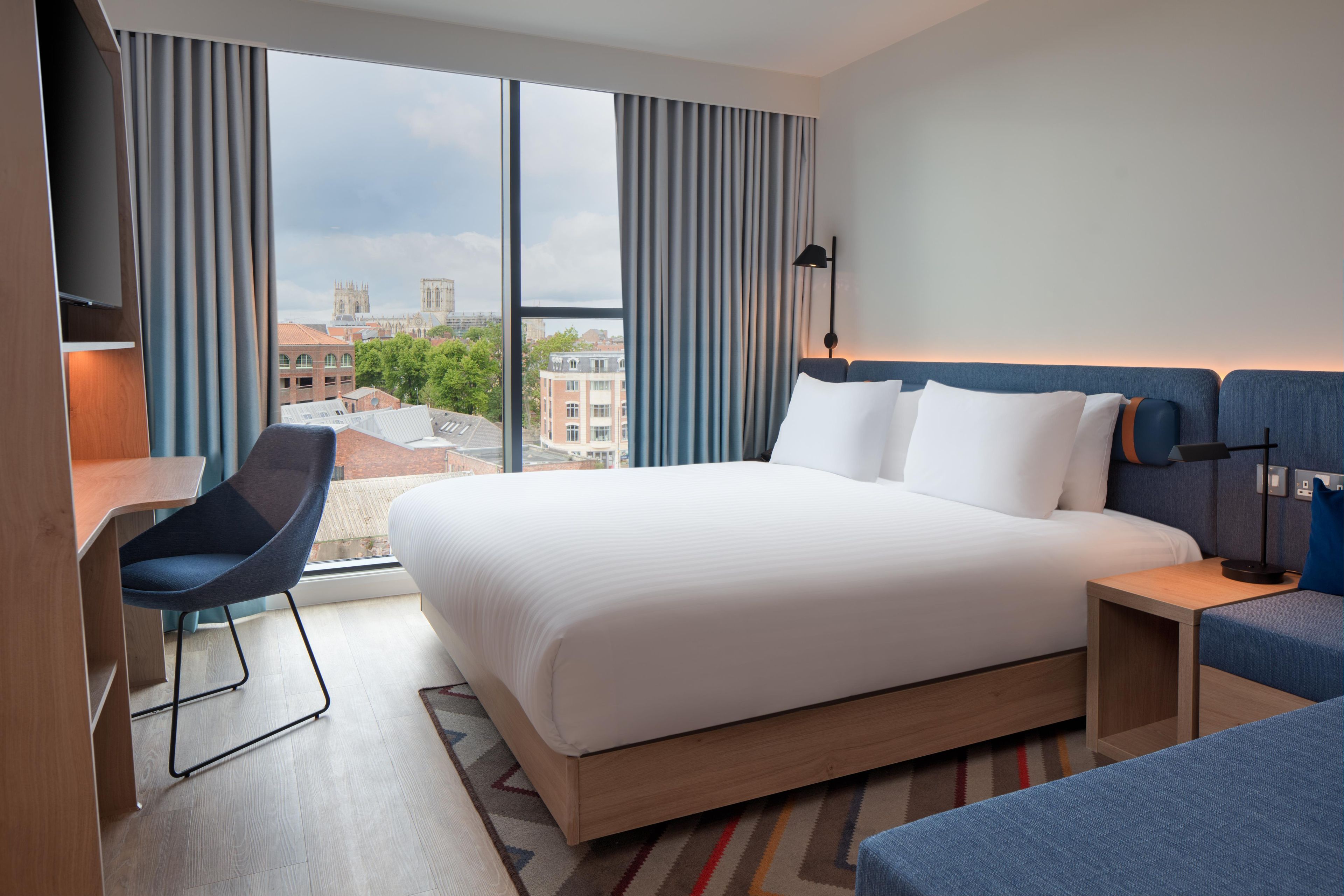 A hotel room with a large bed, a desk, a chair, and a view of the cityscape.