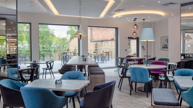 A modern dining area with multiple tables and chairs, large windows offering a view of the outdoors, and a mix of pendant and table lamps providing lighting.