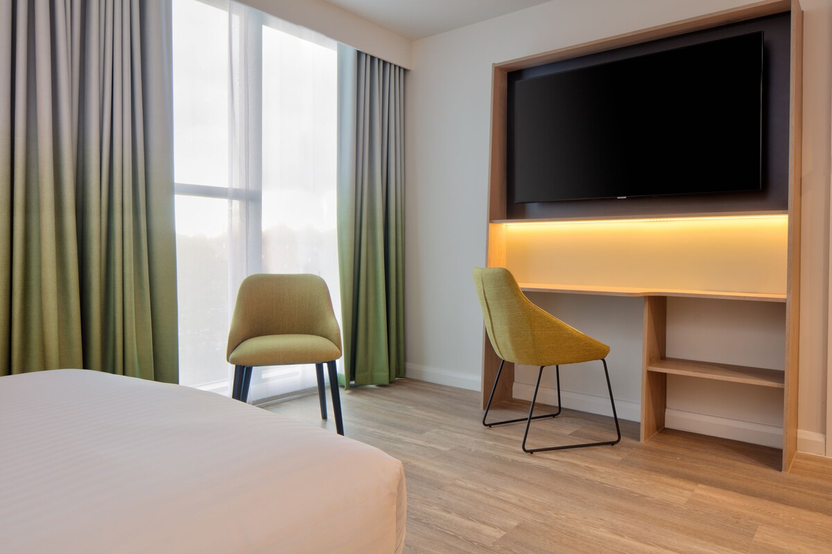 A hotel room with a large bed, two chairs, a flat-screen TV mounted on the wall, and a window with curtains.