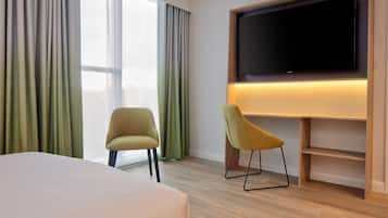 Ein Hotelzimmer mit einem großen Bett, zwei Stühlen, einem flachbildfernseher an der Wand und einem fenster mit Vorhängen.
