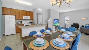 Eine moderne Küche mit Holzausstattung, einem weißen Kühlschrank und einer Mikrowelle. Ein Essbereich mit Tisch für sechs Personen, blauen Stühlen und gewebten Tischsets. Eine Kristallleuchter hängt über dem Esstisch.