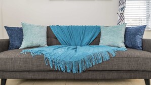 Ein graues Sofa mit einem blauen Überwurf und Fransen, das von zwei Kissen, einem hellblauen und einem dunkelblauen, flankiert wird.
