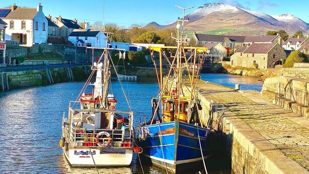 Un puerto con barcos amarrados a un muelle de piedra, rodeado de casas y una montaña al fondo.