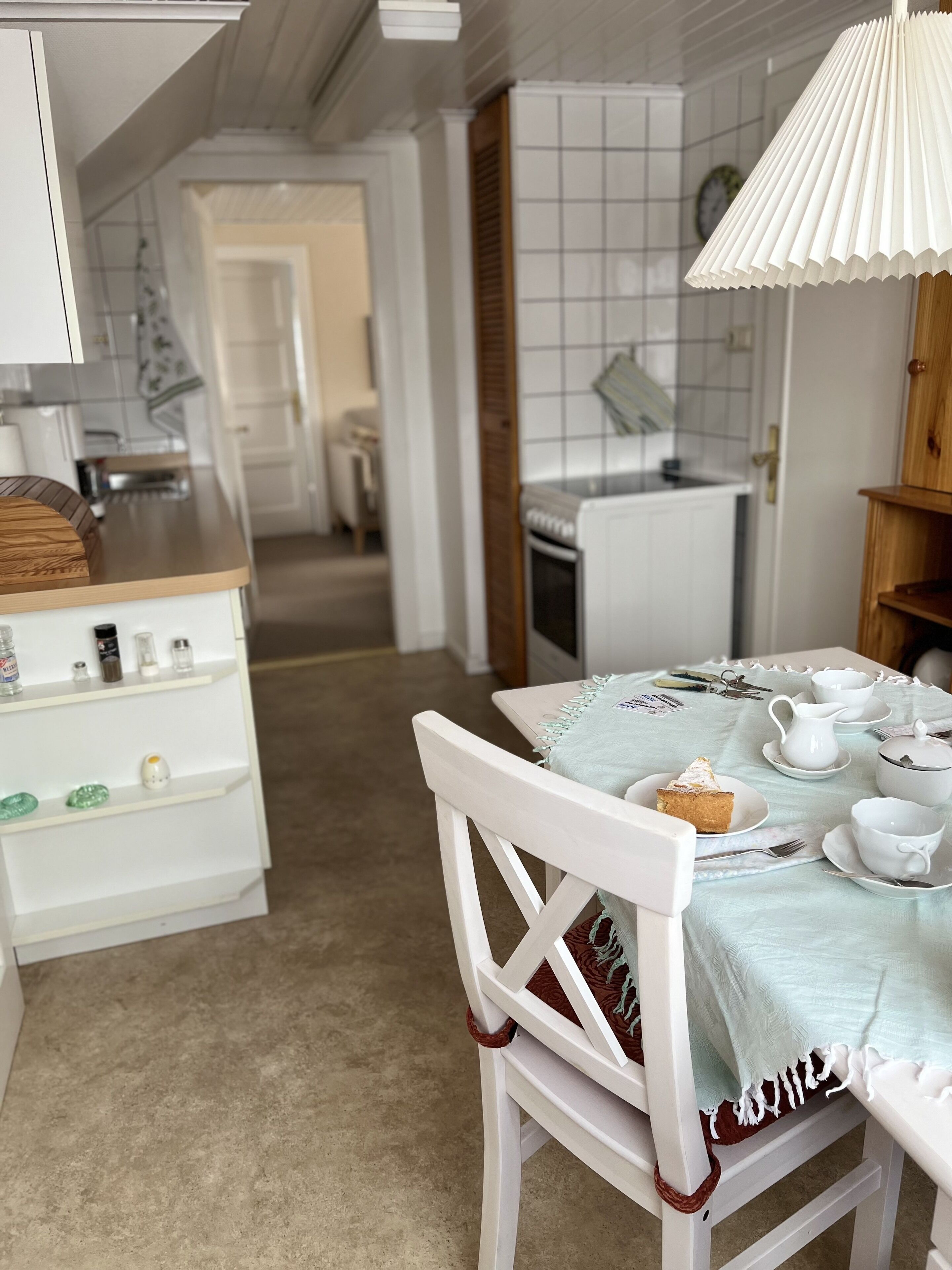 A dining area with a table set for tea, a white chair, and a kitchen in the background.