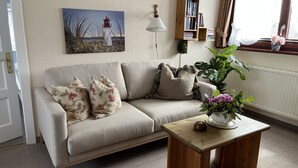 Een woonkamer met een beige bank, bloemenkussens, een houten bijzettafeltje met een bloevas, een boekenkast en een schilderij van een vuurtoren.
