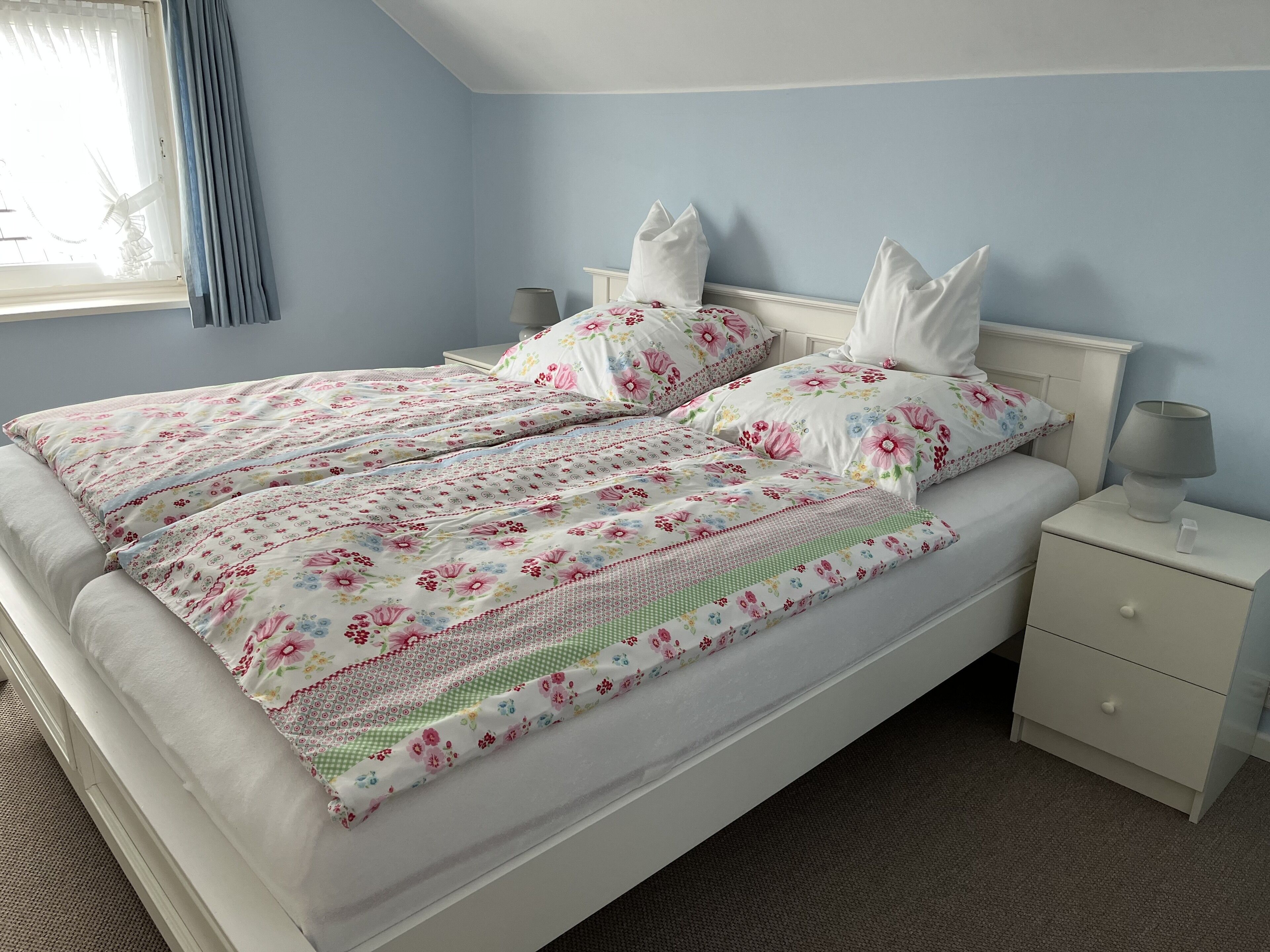 Una cama bien hecha con ropa de cama y almohadas estampadas, una mesita de noche con lámpara y una ventana con cortinas.