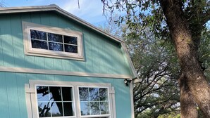 Una casa de dos pisos con fachada color turquesa, dos ventanas y una terraza de madera con sillas Adirondack.