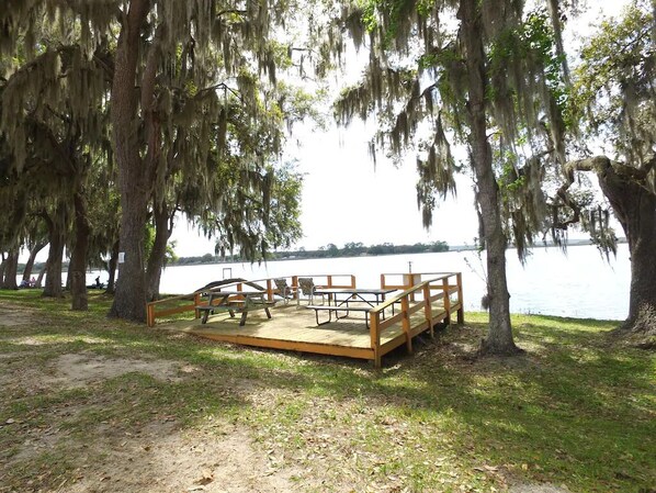 A wooden deck with benches and picnic tables by a lake, surrounded by trees with hanging moss.