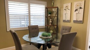 Un comedor con una mesa de vidrio rodeada por cuatro sillas, un estante decorativo con plantas y dos obras de arte enmarcadas en la pared.
