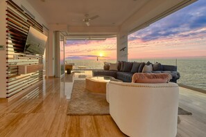 Un salón moderno con un gran ventanal que ofrece vistas a la puesta de sol, un sofá modular y un separador de vidrio con vistas a la ciudad.