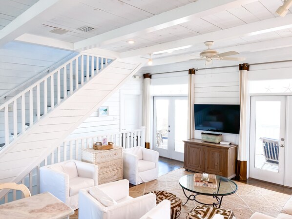 A spacious living room with a white staircase, a glass coffee table, and a large flat-screen TV. - BeachFront House, Ocean View, Pool, 5 Porches, Sleeps 13, Bikes (Santa Rosa Beach)