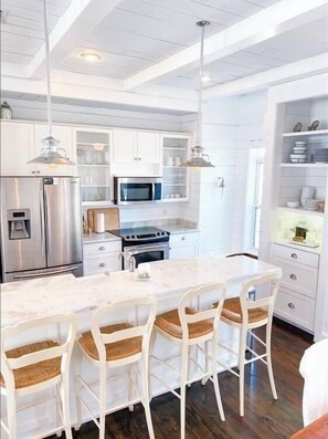 Una cocina moderna con armarios blancos, electrodomésticos de acero inoxidable, una isla de mármol e iluminación colgante.