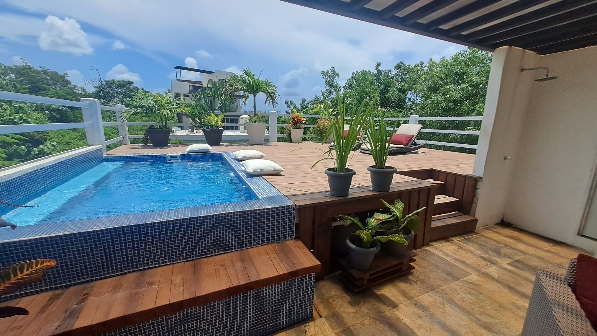 A rooftop pool area with a wooden deck, potted plants, and a view of surrounding greenery and buildings.