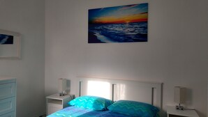 Un dormitorio con cama, mesitas de noche y un cuadro en la pared que muestra un atardecer sobre el océano.