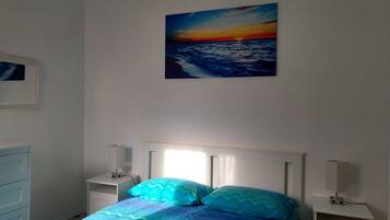 Ein Schlafzimmer mit einem Bett, Nachttischen und einem Wandbild, das einen Sonnenuntergang über dem Meer zeigt.