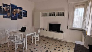 Una habitación pequeña y limpia con una mesa de comedor, sillas, un televisor y una estantería con botellas.