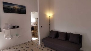 Una habitación pequeña y bien iluminada con un sofá de color oscuro, un estante blanco con utensilios de cocina y un cuadro de un muelle en la pared.