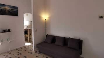 A small, well-lit room with a dark-colored sofa, a white shelf with kitchen items, and a painting of a pier on the wall.