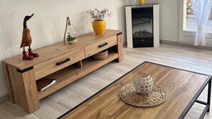 A modern living room with a wooden coffee table, a flat-screen TV mounted on the wall, a wooden cabinet with drawers, a small fireplace, and a window with curtains. - Comfortable house, seaside + Private Parking (Gruissan)