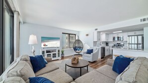 En moderne stue med sofa, sofabord og en hængestol. Der er et køkken med hvide skabe og et spisebord. Et stort vindue giver udsigt til stranden.