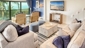Ein geräumiges Wohnzimmer mit einer großen Couch, zwei Sesseln, einem Couchtisch und einem an der Wand montierten Fernseher. Ein Balkon bietet Blick auf den Ozean.
