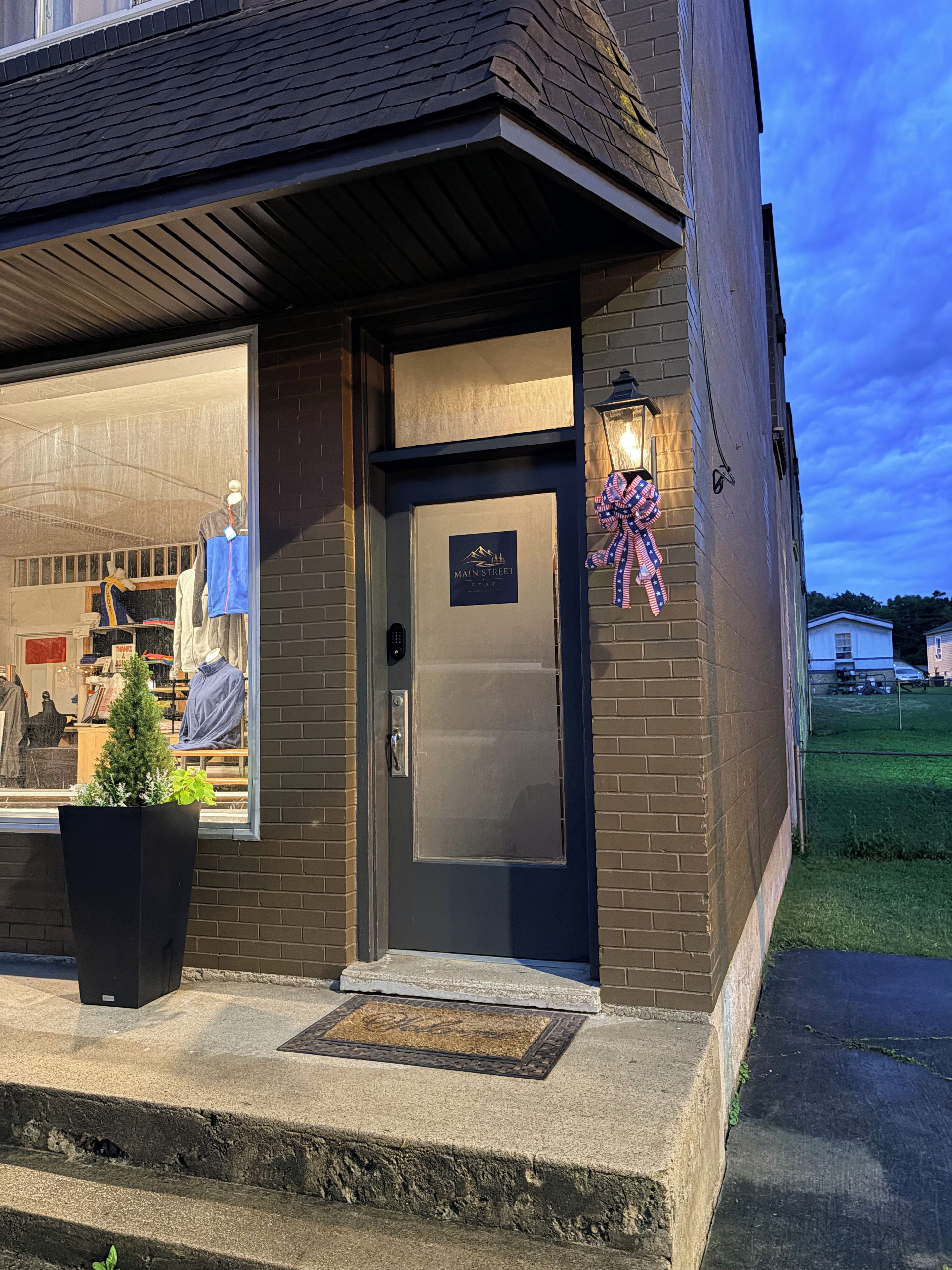 A storefront with a dark brick facade, a clear glass window displaying clothing, a small potted plant, and a door with a sign.