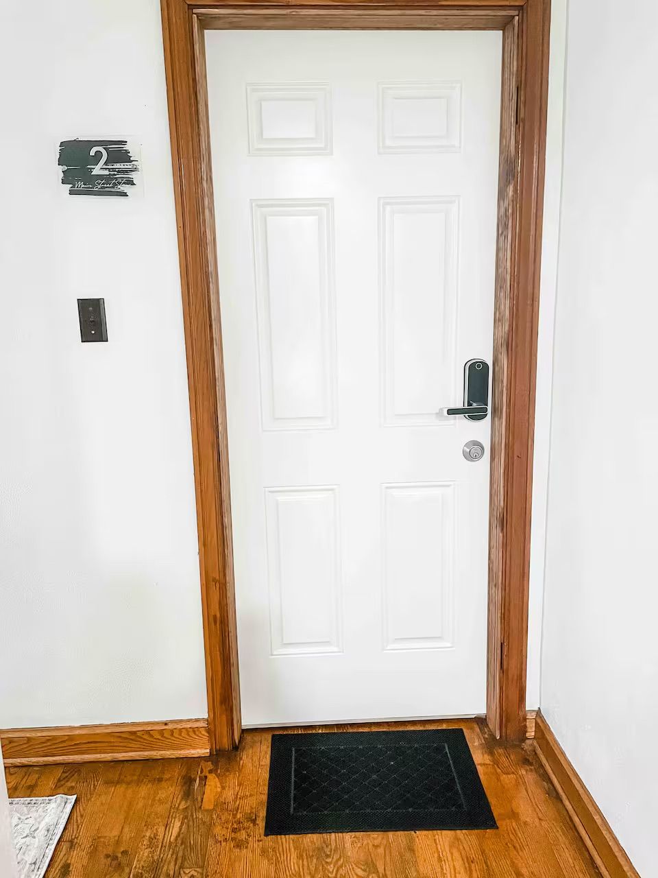 Een witte deur met houten frame, een zwarte deurmat en een klein bord aan de muur.