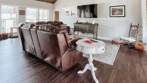 A spacious living room with a high ceiling, wooden beams, a brown leather sofa, a white coffee table, and a television mounted on the wall.