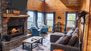 L’intérieur d’un chalet chaleureux, avec une cheminée en pierre, des fauteuils à revêtement bleu, un canapé gris et un plafond en bois.