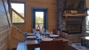 Un intérieur en bois avec une table à manger dressée pour quatre personnes, une cheminée en pierre et une vue sur l’extérieur grâce à une grande fenêtre.