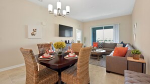 A dining area with a dark wooden table, wicker chairs, and a chandelier. There is a sofa and a television in the background. - Water Views, Premium Resort Amenities Included. (Kissimmee)