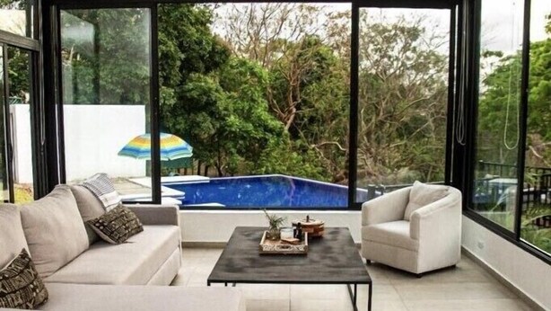 A modern living room with a beige sofa, a coffee table, and a white armchair. A large window offers a view of a swimming pool and greenery. - Casa completa dentro de un complejo privado con seguridad 24/7 (La Libertad)