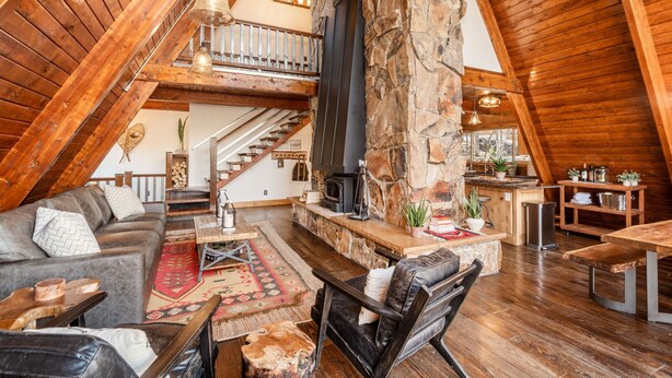 A cozy loft-style living room with a stone fireplace, wooden beams, and a staircase leading to an upper level.