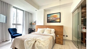 A modern hotel room with a large bed, a desk, a chair, a TV, and a glass door closet.