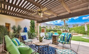 A covered outdoor seating area with green cushioned chairs, a round table, and a view of a swimming pool and mountains.