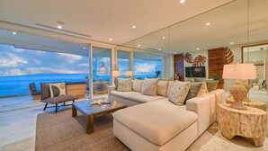 Una sala moderna con un sofá grande, una mesa de centro y un televisor. Hay ventanas amplias que dan a una vista del océano.