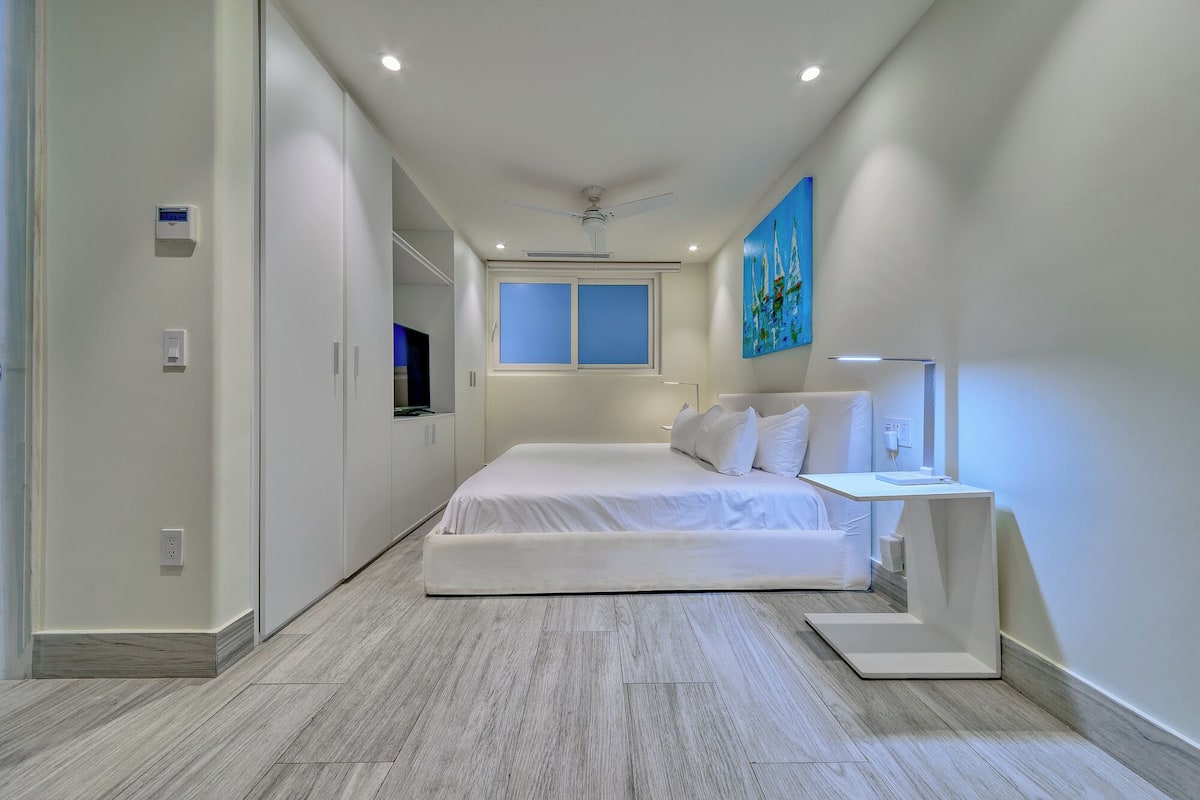 A modern hotel room with a bed, bedside table, ceiling fan, and a window with blue-tinted glass.