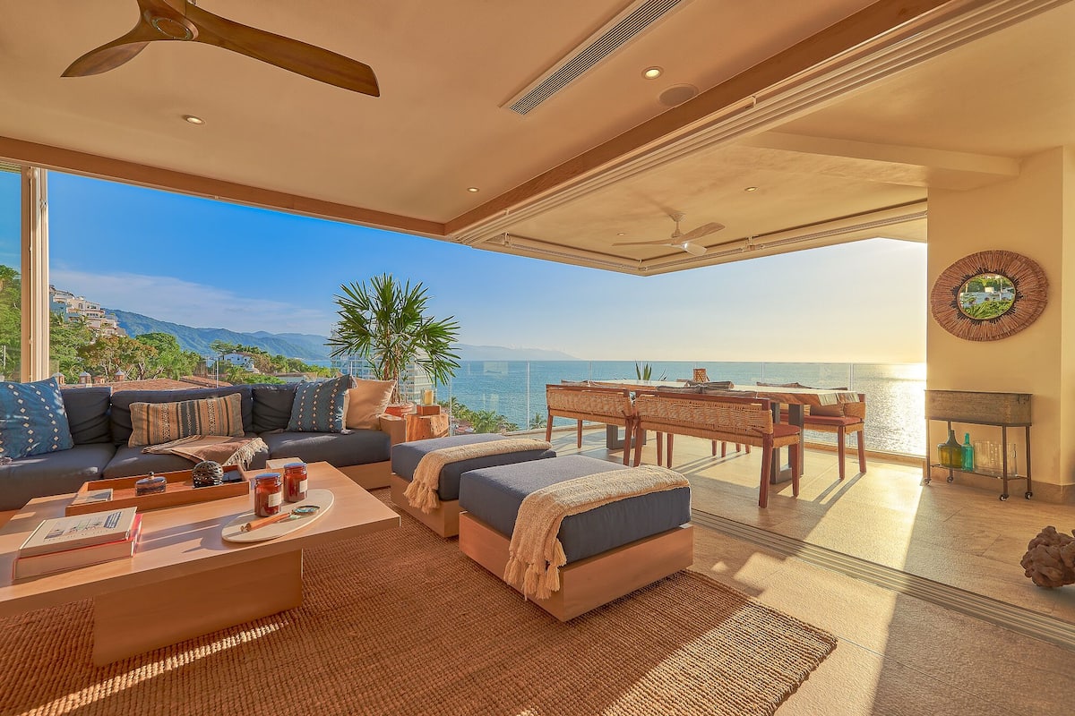 A spacious living area with a large window overlooking a scenic ocean view, furnished with a sofa, coffee table, and a dining table with chairs.