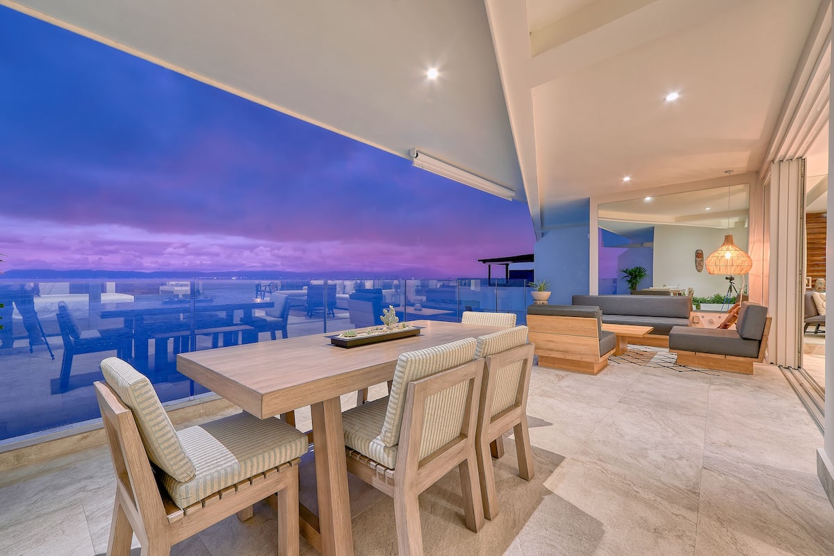 A modern living room with a large window overlooking a cityscape, a wooden dining table with chairs, and a cozy seating area with a sofa and side tables.