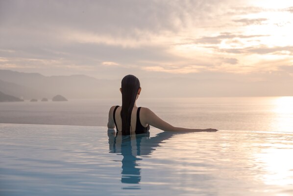 Uma pessoa sentada em uma piscina infinita transparente com vista para o oceano e montanhas ao longe.