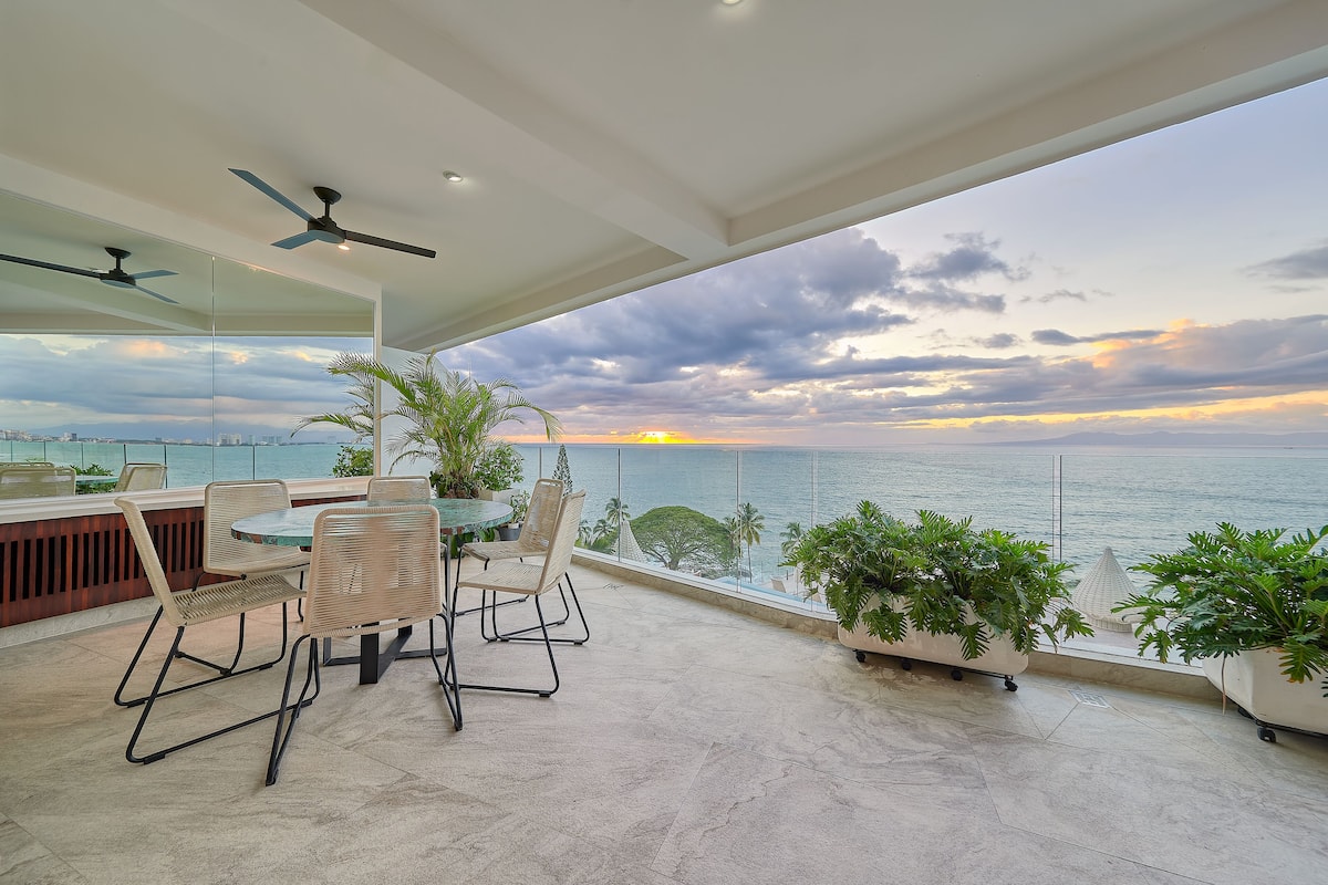 A balcony with a table and chairs, overlooking a body of water and cityscape at sunset.