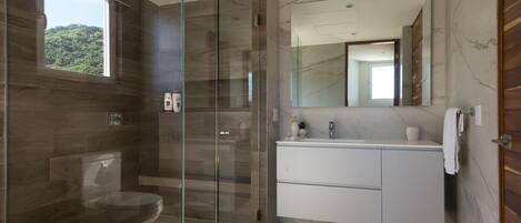A modern bathroom with a glass-enclosed shower, a toilet, a vanity with a mirror, and a view of greenery outside.