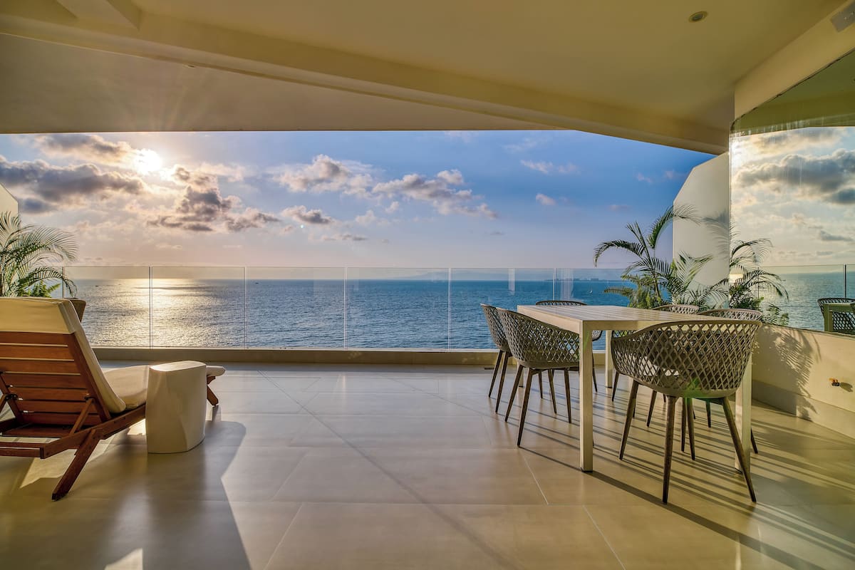 A balcony with a view of the ocean, a wooden chair, a table with chairs, and potted plants.