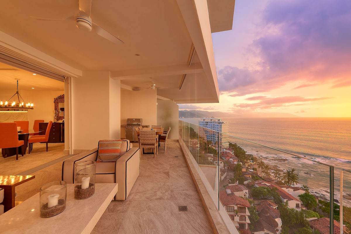 A balcony with a view of the ocean at sunset, a dining area with orange chairs, and a modern kitchen.