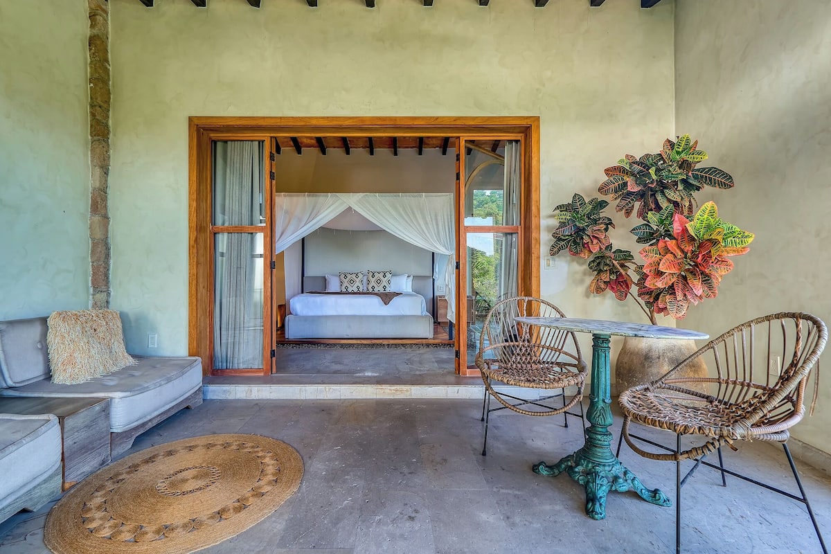 A bedroom with a bed, a sitting area with a sofa and chairs, a small table, and a large potted plant.
