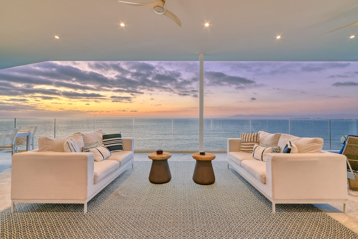 A modern living room with two white sofas, a patterned rug, and a view of the sunset over the ocean.