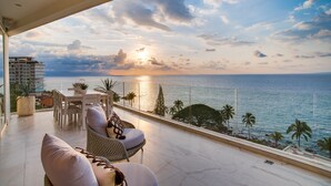 Un balcón con vista al mar, una zona de comedor con sillas y una mesa, y un edificio al fondo.
