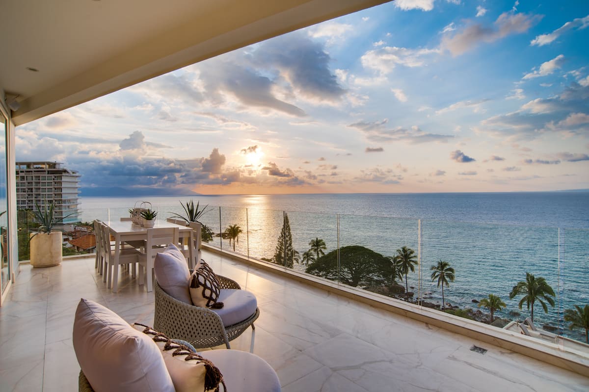 A balcony with a view of the sea, a dining area with chairs and a table, and a building in the background.