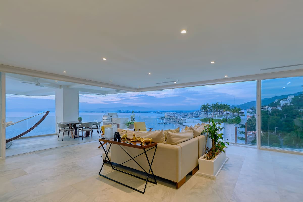 A spacious living room with a large window offering a city and water view, a comfortable sofa, a dining area, and a hammock.